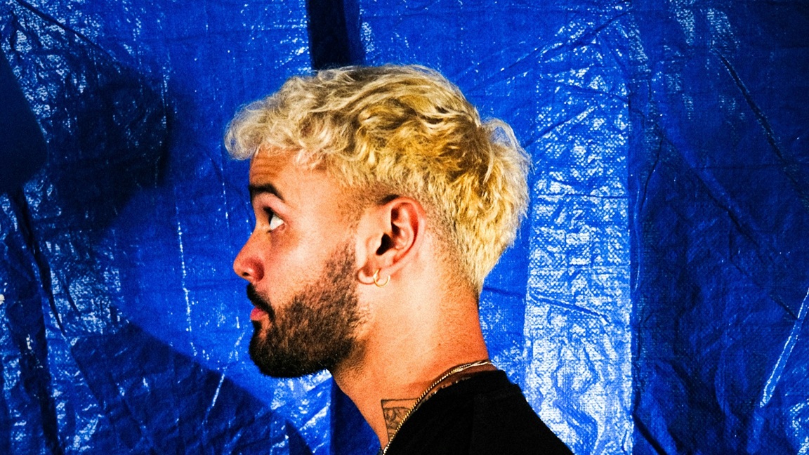 Man met geblondeerd krullend haar en baard, gefotografeerd in profiel voor een blauwe achtergrond &ndash; vet haar