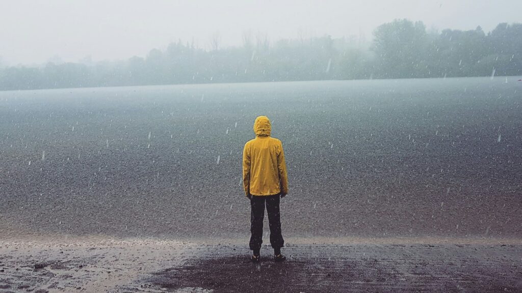 Persoon in gele jas bij regenachtig meer illustreert hoe je lichaamswarmte verliezen kunt door natte en koude omstandigheden
